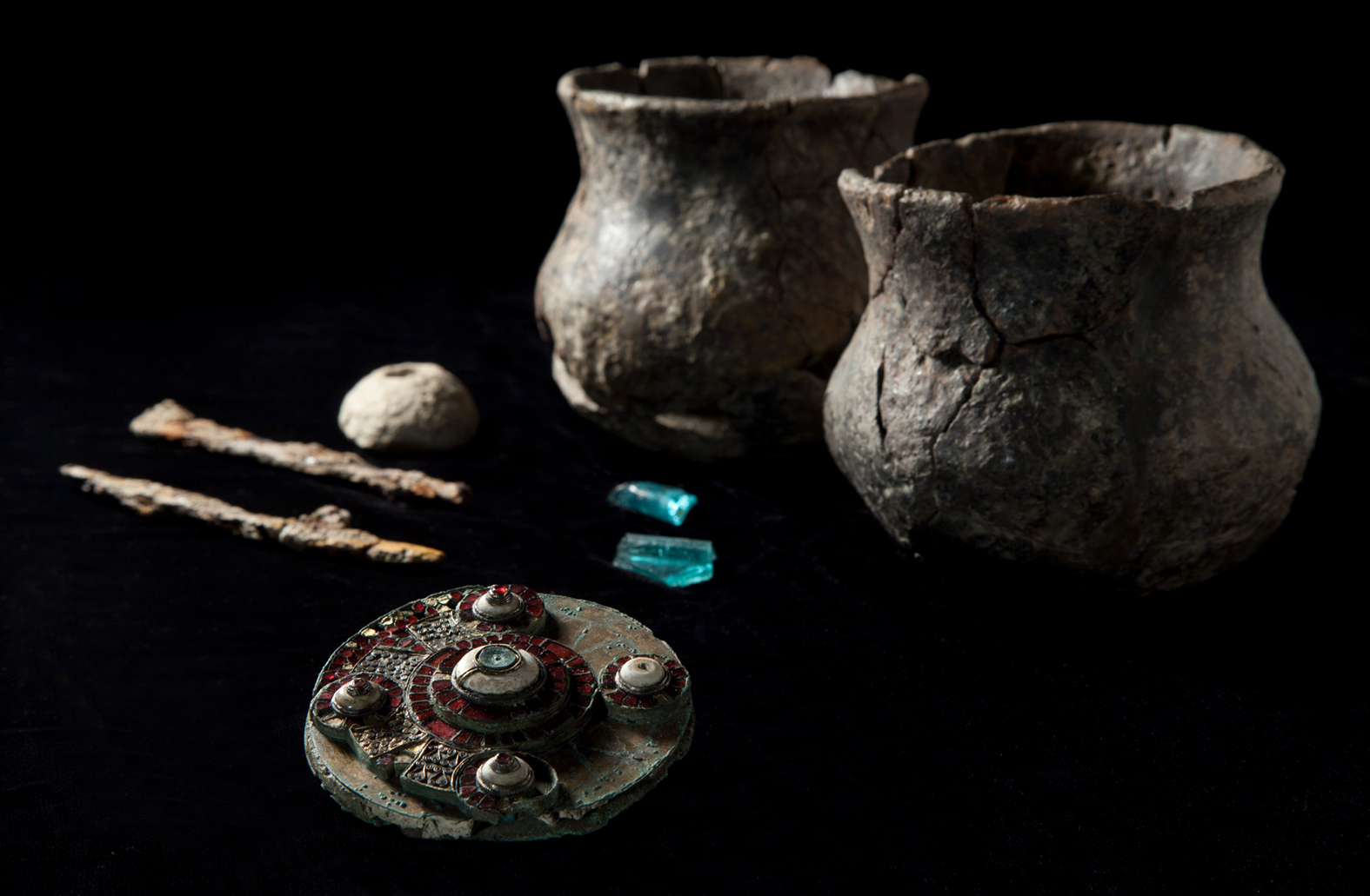 A photo of the burial assemblage discovered at West Hanney, including two fragments of glass (apparently from the same vessel), a chalk and spindle whorl, iron knives,and, two small ceramic vessels (most likely cups). 