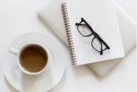 Photo of a table with a notepad, reading glasses and a cup of coffee